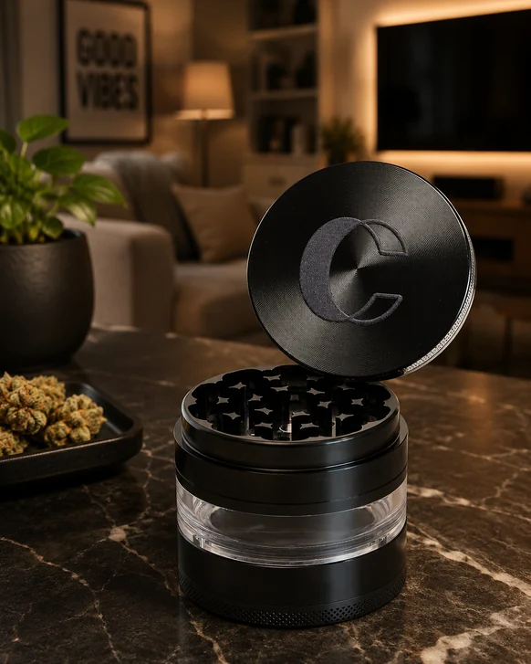 A black four-piece metal herb grinder on a marble countertop, lid tilted off to show the diamond teeth, with a clear window band around the collection chamber