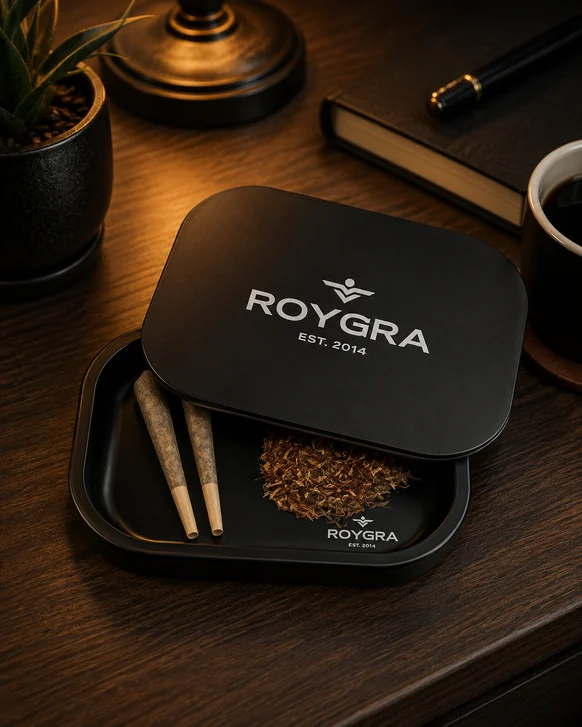 A black ROYGRA rolling tray on a dark wood desk with its magnetic lid slid half open, two cones and a small pile of loose herb inside